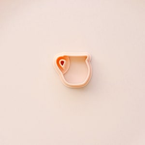 May include: A light peach-colored cookie cutter in the shape of a pitcher. The cutter has a handle and a spout, and the interior is hollow. The cutter is made of plastic and is set against a matching peach background.