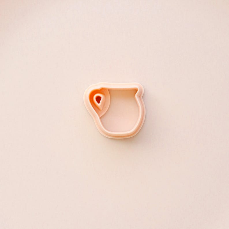 May include: A light peach-colored cookie cutter in the shape of a pitcher. The cutter has a handle and a spout, and the interior is hollow. The cutter is made of plastic and is set against a matching peach background.