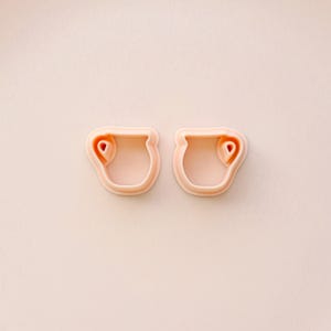 May include: Two light pink cookie cutters in the shape of a small vase or pot with a heart-shaped detail. The cutters are made of plastic and are set against a light pink background.
