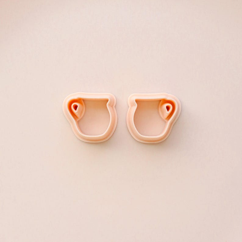 May include: Two light pink cookie cutters in the shape of a small vase or pot with a heart-shaped detail. The cutters are made of plastic and are set against a light pink background.