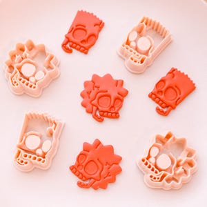 May include: A collection of orange and light pink plastic cookie cutters in the shape of a cartoon skull. The cutters are arranged on a light pink surface. The image shows both the cutter and the cut-out shapes.
