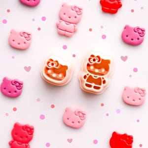 Op de afbeelding: Een verzameling roze en rode Hello Kitty-vormige versieringen verspreid op een witte achtergrond met roze stippen en harten. De versieringen variëren in roze en rode tinten, sommige met een strik.