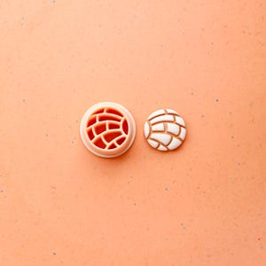 May include: A light peach-colored cookie cutter and a concha cookie. The cutter is round with a brick-like pattern. The concha cookie is white with a brown base, also with a brick-like pattern.