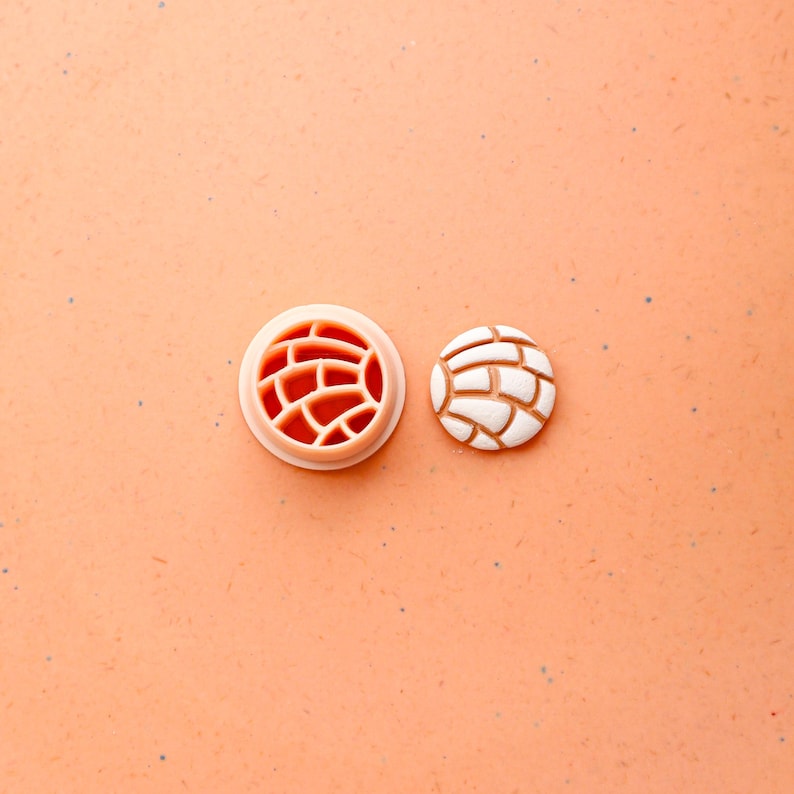 May include: A light peach-colored cookie cutter and a concha cookie. The cutter is round with a brick-like pattern. The concha cookie is white with a brown base, also with a brick-like pattern.