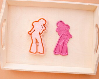 May include: Two cookie cutters in the shape of a woman in a disco outfit. One is light orange, the other is pink. The cookie cutters are on a wooden tray with a light orange background. The woman is wearing flared pants and a top.