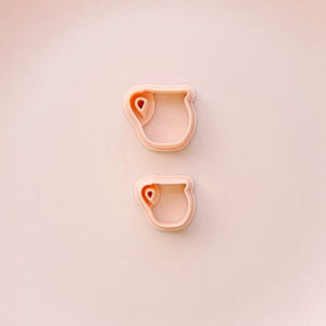 May include: Two light pink cookie cutters in the shape of a teacup with a handle. The cutters are made of plastic and are set against a light pink background. The teacup design includes a small, rounded handle and a curved rim.