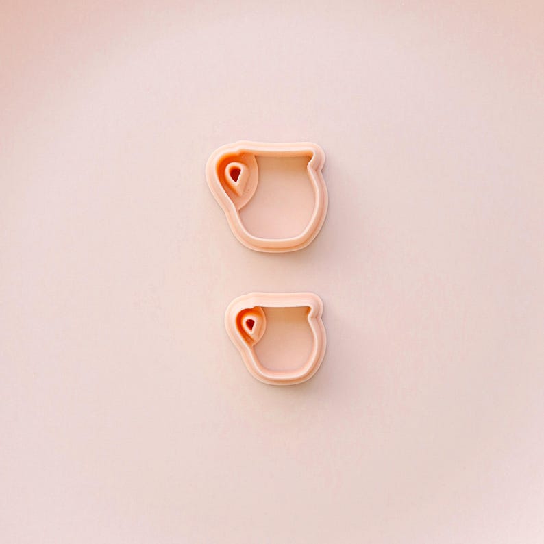 May include: Two light pink cookie cutters in the shape of a teacup with a handle. The cutters are made of plastic and are set against a light pink background. The teacup design includes a small, rounded handle and a curved rim.