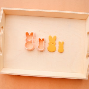 May include: Four bunny-shaped cookie cutters in a wooden tray. Two are pink plastic cutters, and two are yellow, with the shapes of the Easter candy. The tray is light brown, and the background is peach.