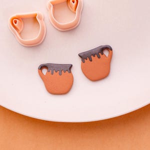 Op de afbeelding: Een close-up van een witte plaat met twee bruine keramische mokken met donkerbruine bovenkanten en handvatten. Twee roze plastic mokvormige koekjessnijders zijn ook zichtbaar. De mokken lijken versierd met een donkerbruin glazuur.