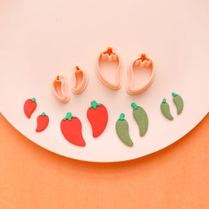 May include: A collection of red and green chili pepper-shaped cookie cutters and matching edible decorations. The cutters are light pink, and the peppers are arranged on a white plate against an orange background. The peppers vary in size.
