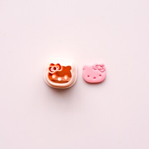 May include: Two Hello Kitty-themed items on a pink background. One is a cookie cutter with a white frame and an orange interior. The other is a pink, die-cut Hello Kitty face with a bow.