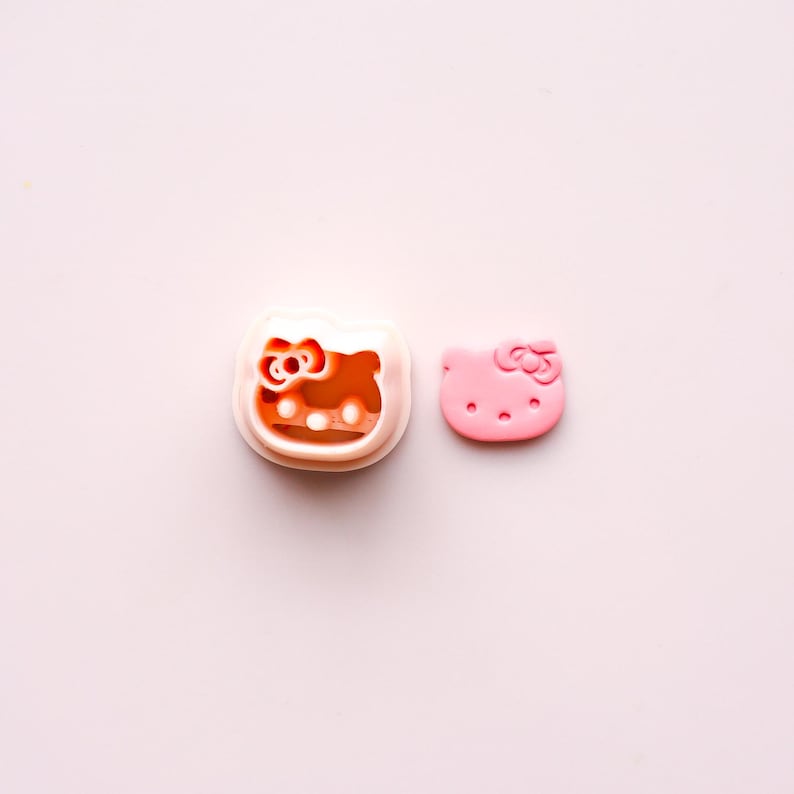 May include: Two Hello Kitty-themed items on a pink background. One is a cookie cutter with a white frame and an orange interior. The other is a pink, die-cut Hello Kitty face with a bow.