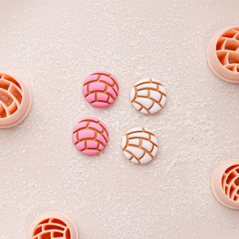 May include: Overhead shot of concha-shaped cookies, some pink and some white, with brown grid patterns. The cookies are arranged on a light pink surface dusted with white powder, with several cookie cutters visible.