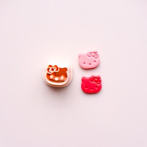 May include: A white and orange Hello Kitty face cookie cutter next to two Hello Kitty face-shaped decorations. One decoration is light pink, and the other is red. All items are on a light pink background.