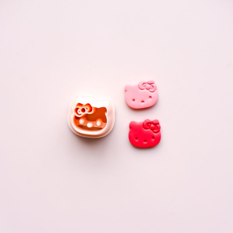 May include: A white and orange Hello Kitty face cookie cutter next to two Hello Kitty face-shaped decorations. One decoration is light pink, and the other is red. All items are on a light pink background.