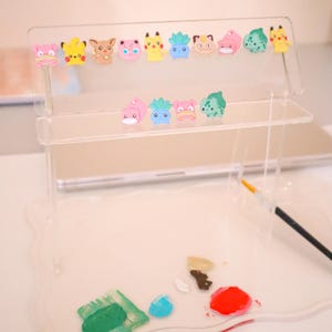 May include: A clear acrylic display stand featuring colorful cartoon character charms. The stand has two shelves and a scalloped base, with a paintbrush and paint splotches in the foreground. The charms include various Pokemon characters in pink, yellow, and green.