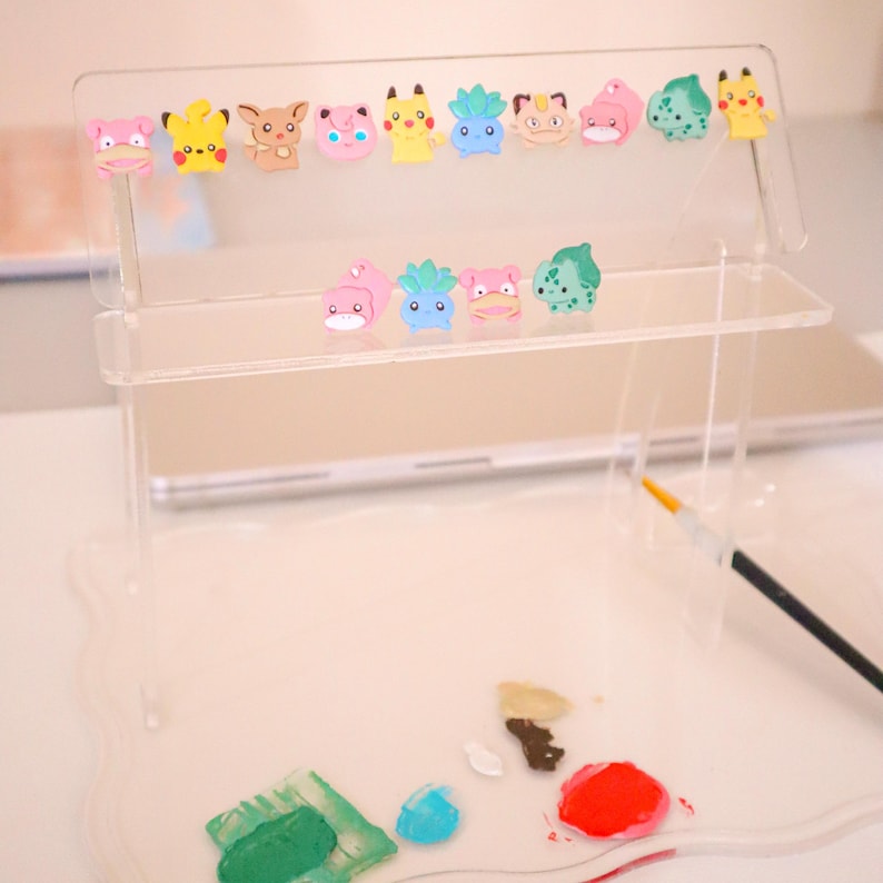 May include: A clear acrylic display stand featuring colorful cartoon character charms. The stand has two shelves and a scalloped base, with a paintbrush and paint splotches in the foreground. The charms include various Pokemon characters in pink, yellow, and green.