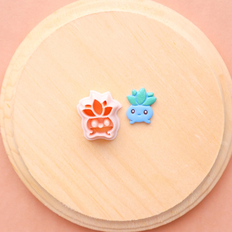 May include: Two colorful cookie cutters shaped like cartoon plants. One is light pink with orange details, and the other is blue with green leaves. They are placed on a light wooden surface.
