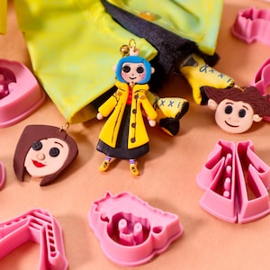 May include: A collection of Coraline-themed items, including a doll charm with blue hair and a yellow coat, face charms, and pink cookie cutters. The doll charm is the focal point, with a detailed design and button eyes.