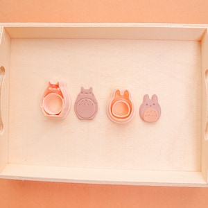 May include: Four pink Totoro-shaped cookie cutters in a wooden tray. The cutters are in various sizes and designs, with detailed features. The tray is light-colored, and the background is a soft peach.
