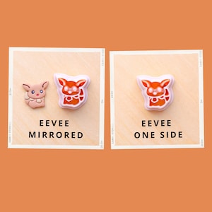 May include: Two Eevee-themed charms, one mirrored and one single-sided, displayed on a light wooden surface. The mirrored charm features a pink and brown design, while the single-sided charm is orange and white. Text reads "EEVEE MIRRORED" and "EEVEE ONE SIDE."