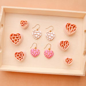 May include: A wooden tray holds heart-shaped earrings and decorative pieces. Two pairs of earrings feature white and pink heart designs with gold-colored hooks. Several heart-shaped pieces in pink are also present, creating a cohesive theme.