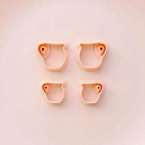 May include: Four light pink cookie cutters in a pitcher shape with a heart detail. The cutters are arranged in a square on a matching pink background. These are ideal for baking and crafting.