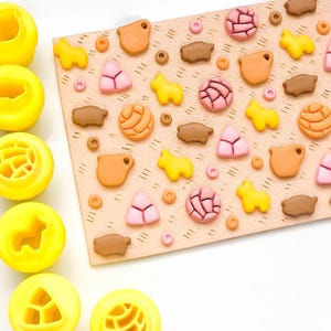 Könnte beinhalten: Eine Sammlung gelber Ausstecher in verschiedenen Formen, darunter ein Kreis, ein Stern und ein Dreieck. Eine rechteckige, beige Oberfläche ist mit kleinen, farbenfrohen Dekorationen zum Thema Essen bedeckt, wie rosa Dreiecken und gelben Tierformen.