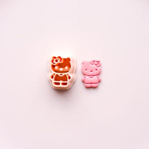 May include: A pink Hello Kitty cookie cutter and a pink Hello Kitty figurine are displayed on a light pink surface. The cookie cutter is a light orange color. The figurine is a solid pink color.