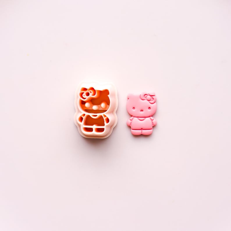 May include: A pink Hello Kitty cookie cutter and a pink Hello Kitty figurine are displayed on a light pink surface. The cookie cutter is a light orange color. The figurine is a solid pink color.