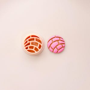 May include: A pink concha cookie cutter and a concha cookie. The cutter is a light pink circle with a brick-like pattern. The cookie is pink with a brown, brick-like design on top. The items are on a light pink surface.