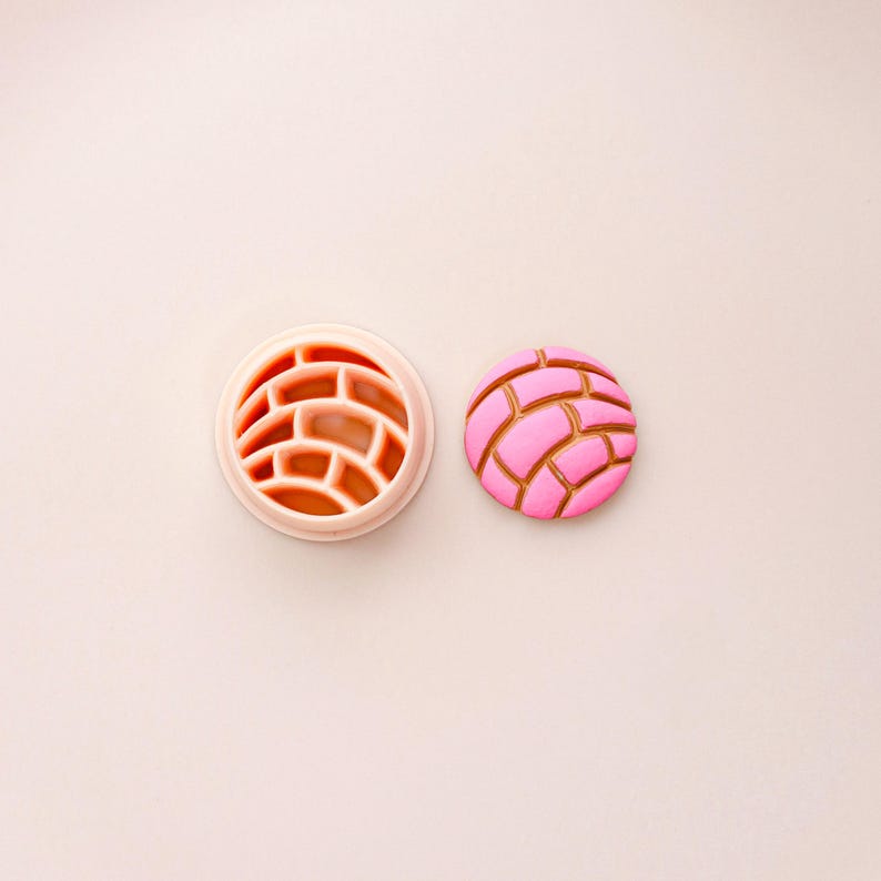 May include: A pink concha cookie cutter and a concha cookie. The cutter is a light pink circle with a brick-like pattern. The cookie is pink with a brown, brick-like design on top. The items are on a light pink surface.