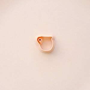 May include: A light peach-colored cookie cutter in the shape of a mug. The cutter has a rounded handle and a small heart-shaped detail inside the mug. The background is a solid light peach color.