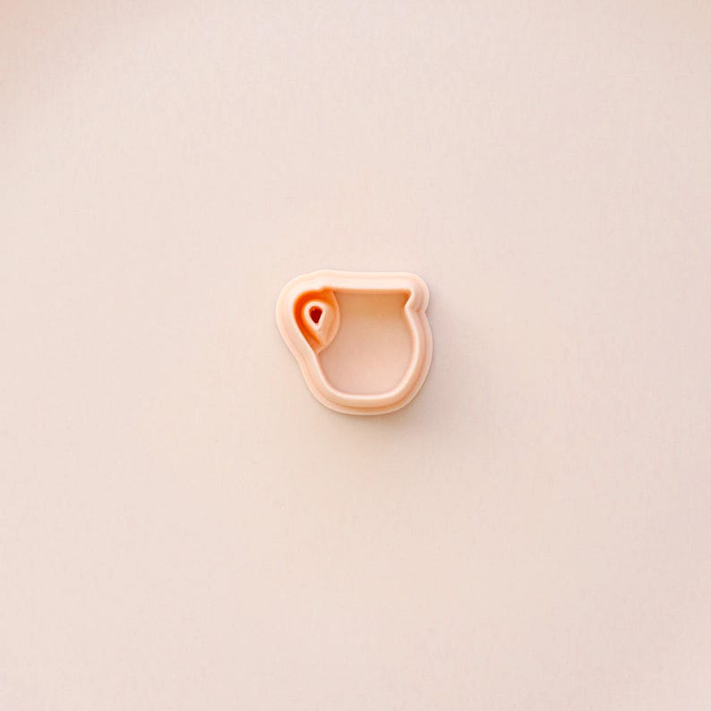 May include: A light peach-colored cookie cutter in the shape of a mug. The cutter has a rounded handle and a small heart-shaped detail inside the mug. The background is a solid light peach color.