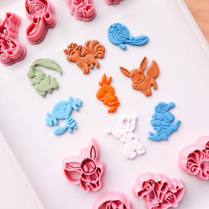 May include: Assorted colorful cookie cutters in the shapes of cartoon characters. The cutters are in shades of pink, orange, blue, green, and white. The cutters are arranged on a white surface.