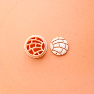 May include: A light orange plastic cookie cutter next to a white and tan cookie. The cutter is round with a brick-like pattern. The cookie is round with a similar brick-like pattern on top. The background is a light orange color.