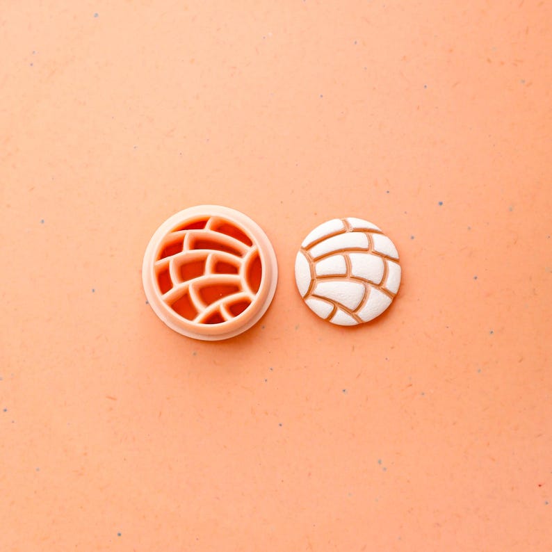 May include: A light orange plastic cookie cutter next to a white and tan cookie. The cutter is round with a brick-like pattern. The cookie is round with a similar brick-like pattern on top. The background is a light orange color.