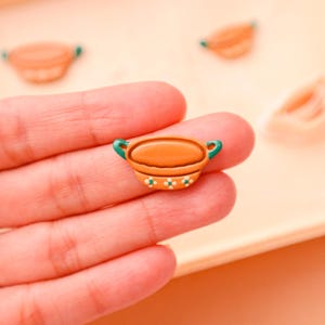 May include: A miniature orange ceramic bowl with green handles and white floral accents. The small bowl is held in a person's hand, with other similar bowls visible in the background. The bowl is likely a decorative item or part of a craft project.