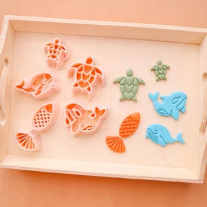 May include: A wooden tray holds a collection of colorful sea creature cookie cutters. The cutters include turtles, whales, and fish in shades of pink, green, orange, and blue. The tray is made of light-colored wood.