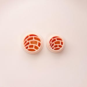 May include: Two round, light pink cookie cutters with a brick pattern design. The cutters are different sizes, with the larger one on the left and the smaller one on the right. The brick pattern is a reddish-orange color.