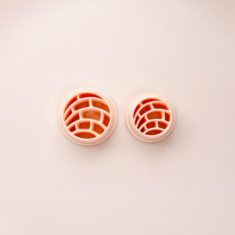 May include: Two round, light pink cookie cutters with a brick pattern design. The cutters are different sizes, with the larger one on the left and the smaller one on the right. The brick pattern is a reddish-orange color.