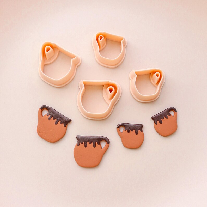 May include: A collection of cookie cutters and cookies shaped like mugs. The cutters are light peach-colored plastic, and the cookies are brown with a dark chocolate-colored topping. The mugs have a handle on the side.