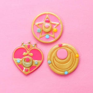 May include: Three pink and gold decorative badges with celestial and heart designs. One badge is a star shape, another is a heart shape with a crown, and the third is a circle with a crescent moon. Each badge has colorful accents.