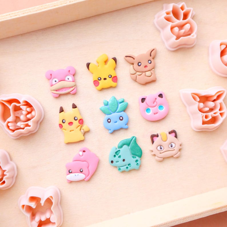 May include: A collection of colorful, cartoon character-shaped cookie cutters and fondant decorations. The designs include various characters in yellow, pink, blue, and brown. The items are arranged on a light wooden surface.