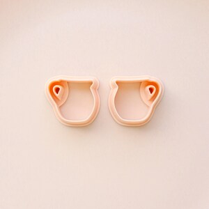 May include: Two light pink cookie cutters in the shape of small pitchers. Each cutter has a rounded body with a handle-like detail on the side. The background is a matching light pink.