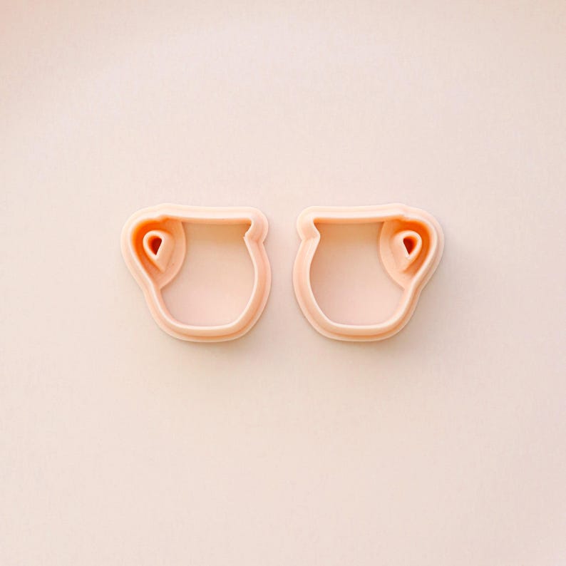May include: Two light pink cookie cutters in the shape of small pitchers. Each cutter has a rounded body with a handle-like detail on the side. The background is a matching light pink.