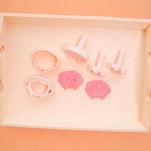 Puede incluir: Un juego de cortadores de galletas de color rosa claro con caras de personajes y formas de cabello. El juego incluye cortadores de cara y cabello, junto con sellos para rasgos faciales. Los cortadores están dispuestos en una bandeja de madera.