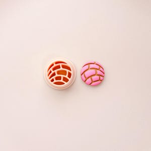 May include: A pink and brown concha cookie cutter and a concha cookie. The cookie cutter is light pink with a brick-like pattern. The cookie is pink with brown lines, mimicking the design of a concha bread roll.