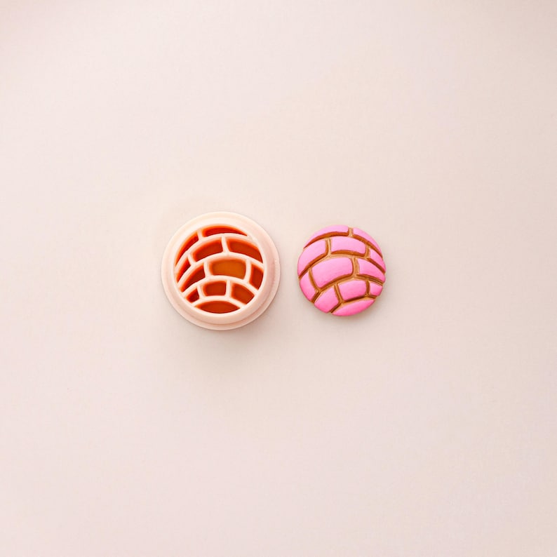 May include: A pink and brown concha cookie cutter and a concha cookie. The cookie cutter is light pink with a brick-like pattern. The cookie is pink with brown lines, mimicking the design of a concha bread roll.
