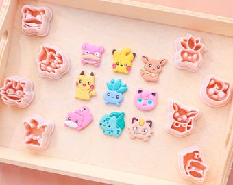 May include: A wooden tray holds an assortment of colorful character-shaped cookie cutters and matching character-shaped cookies. The cookie cutters are pink, and the cookies are yellow, pink, blue, and brown.